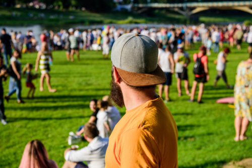 Hipster in cap happy celebrate event fest or festival. Summer fest. Man bearded hipster in front of crowd. Open air concert. Fan zone. Music festival. Entertainment Visit summer festival